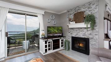 A living room with a fireplace, a television, and a balcony with a view of the mountains.