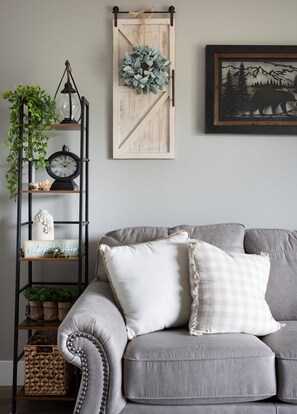 Ein graues Sofa mit dekorativen Kissen, ein an der Wand befestigtes Holzschild mit einem Kranz, ein gerahmtes Bild eines Bären und ein Metallregal mit einer Uhr und Pflanzen.
