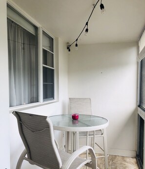 A small, round glass table with a white metal chair, a red candle, and a string of light bulbs on the ceiling. - The Intercostal Condo (Hallandale Beach)