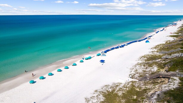 Une plage de sable blanc bordée d’une eau turquoise. De nombreux parasols bleus et des chaises de plage sont alignés le long du rivage.