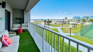 A balcony with a white railing, a white sofa with a pink pillow, and a red pot. There is a view of a pool area with a fence and buildings in the background. - Beach Please 2BR Gulf Shores Oceanfront/Pool, Beach Please Beachfront condo sleeps 6 (Baldwin County)