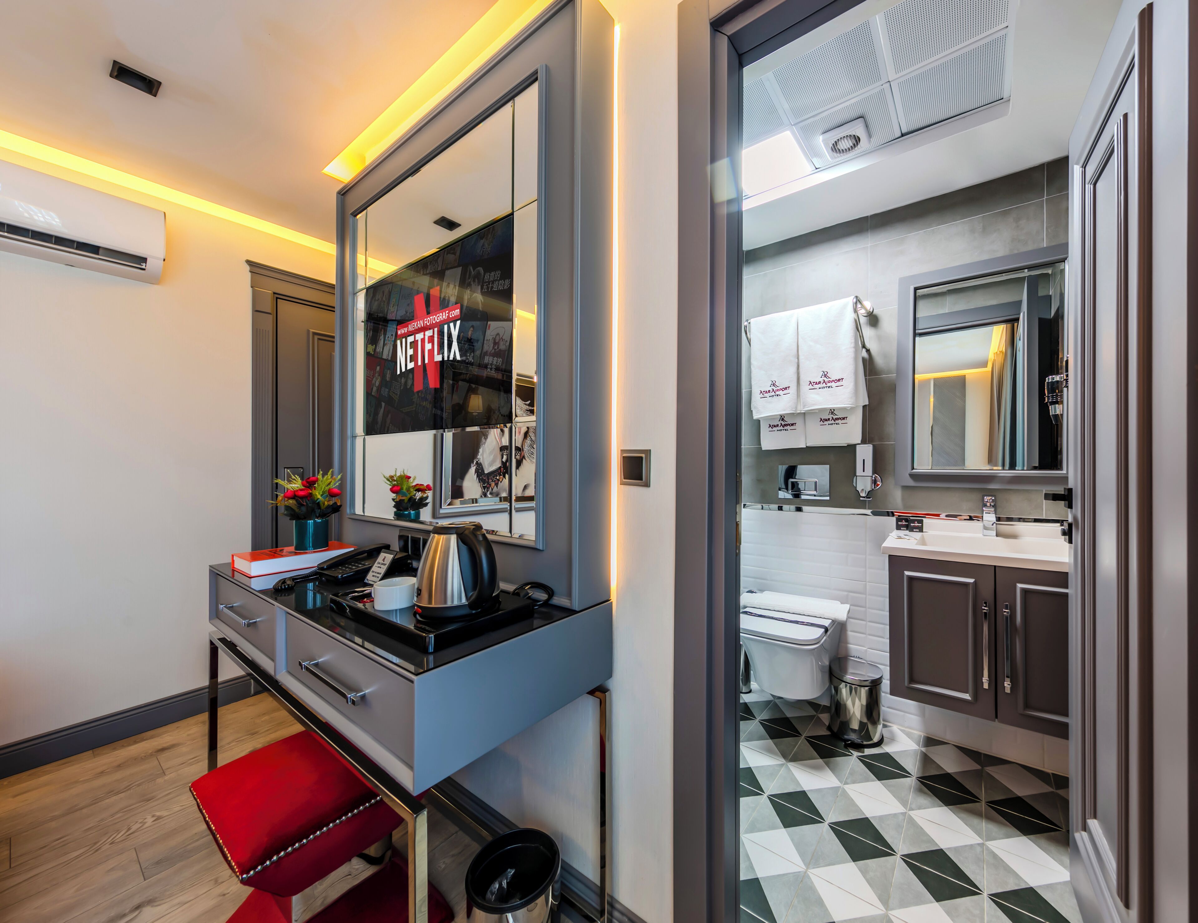 A modern hotel room with a large mirror, a desk with a kettle, a red bench, and a view of the bathroom through a glass door.