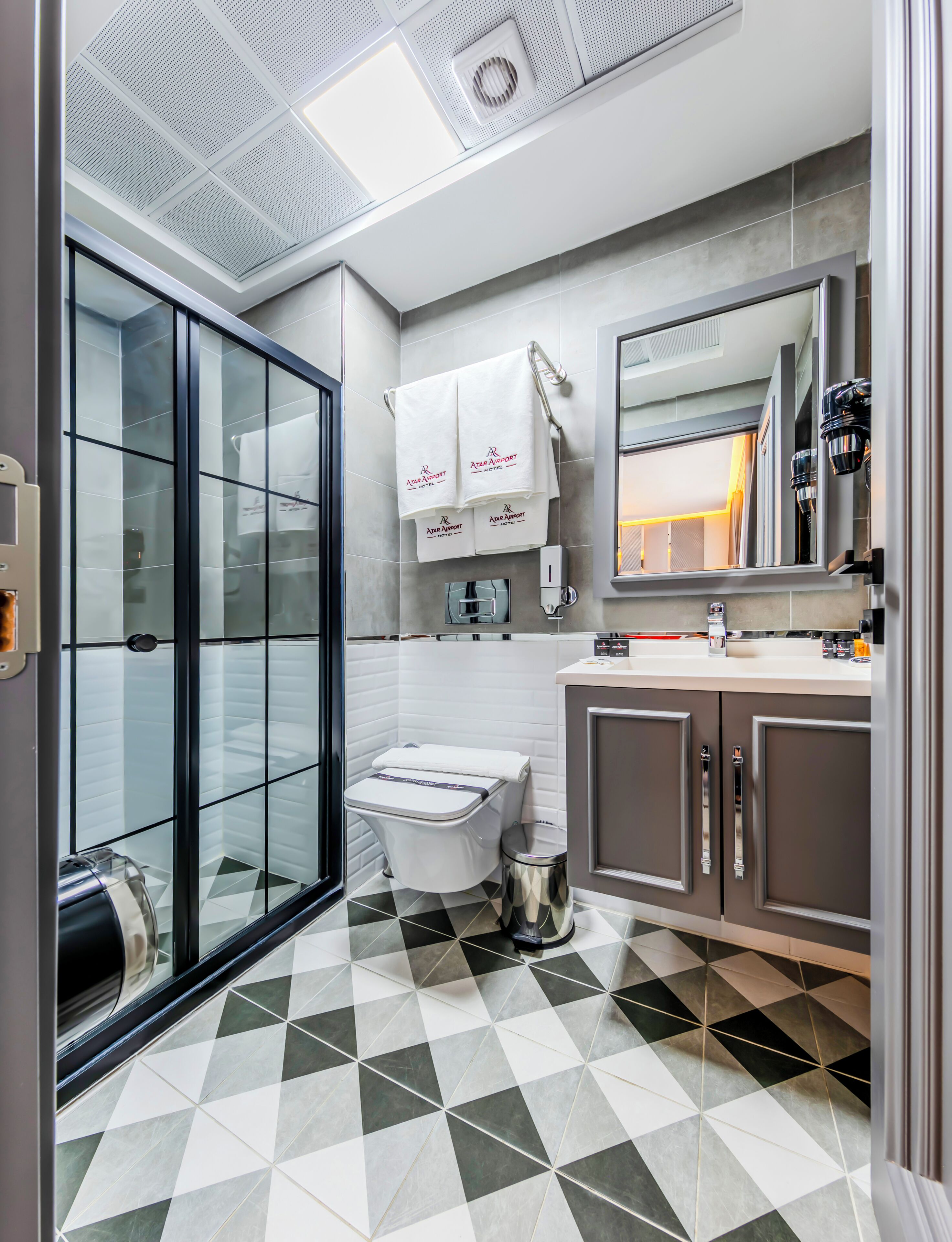 A modern bathroom with a glass shower enclosure, a toilet with a privacy partition, a sink with a mirror, and a towel rack with branded towels.