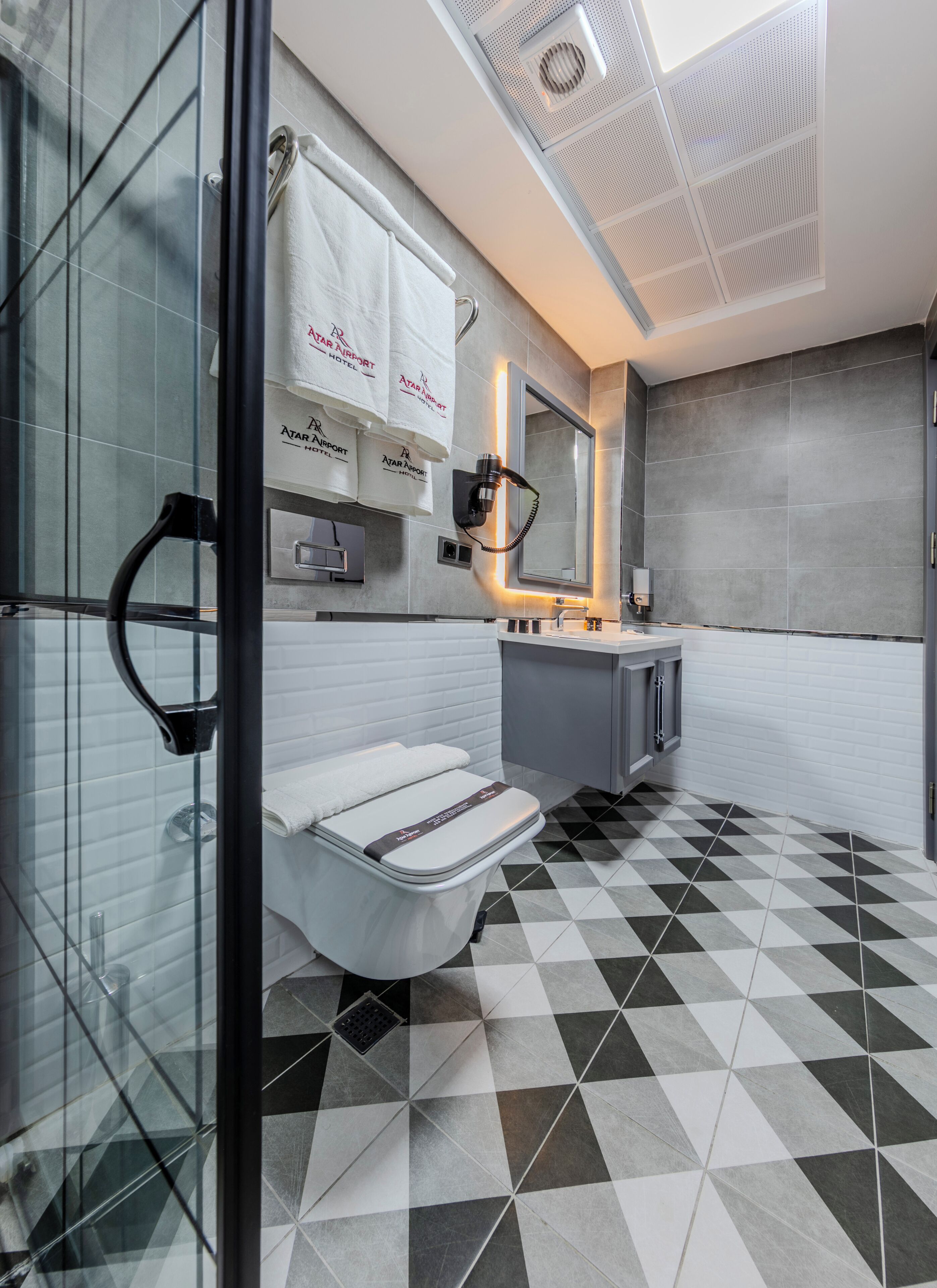 A modern bathroom with a checkered floor, a glass shower enclosure, a vanity with a mirror, and a towel rack with branded towels.
