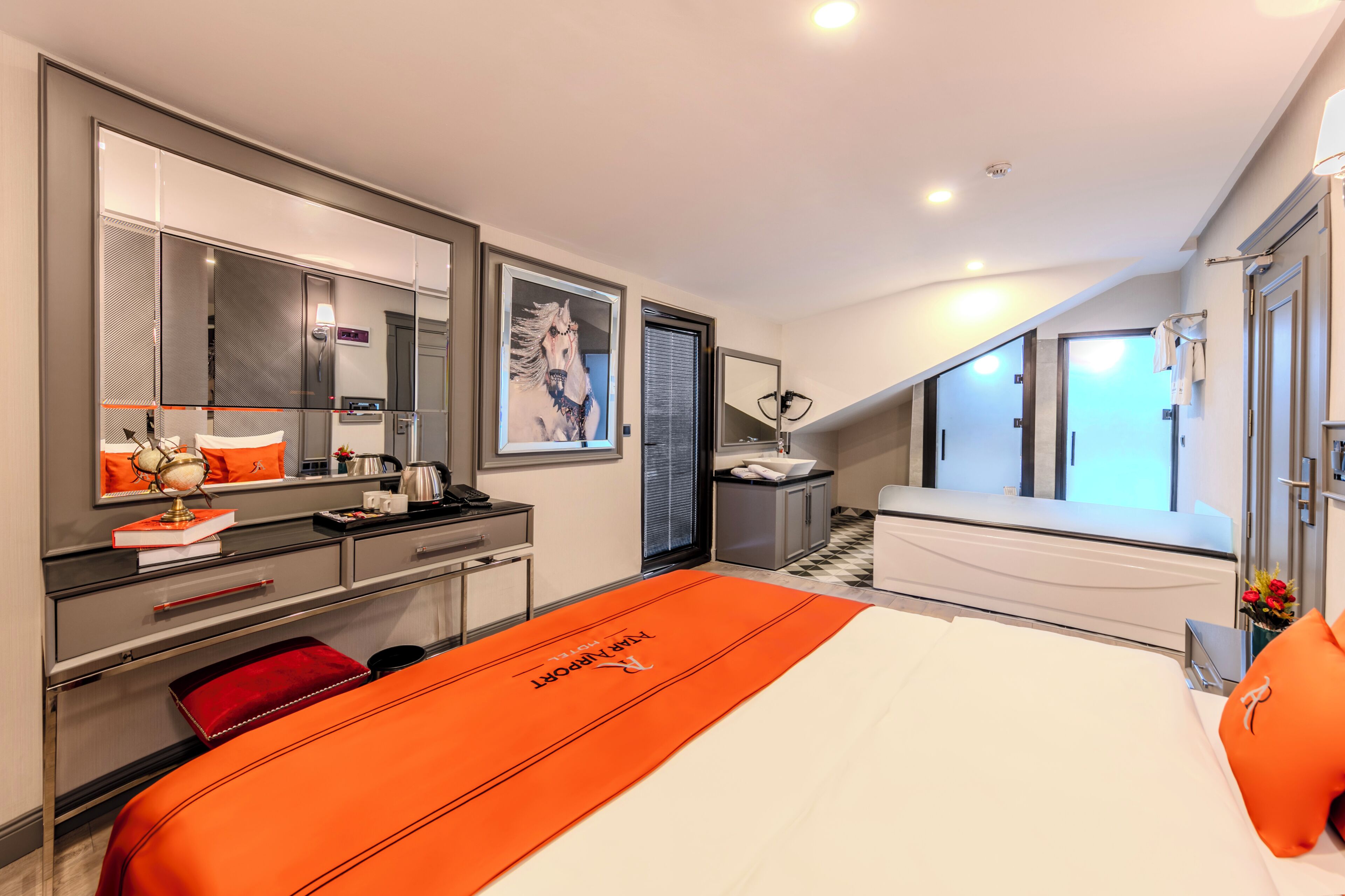A modern hotel room with a large bed, a desk with a computer, a mirror, and a view of the hallway.