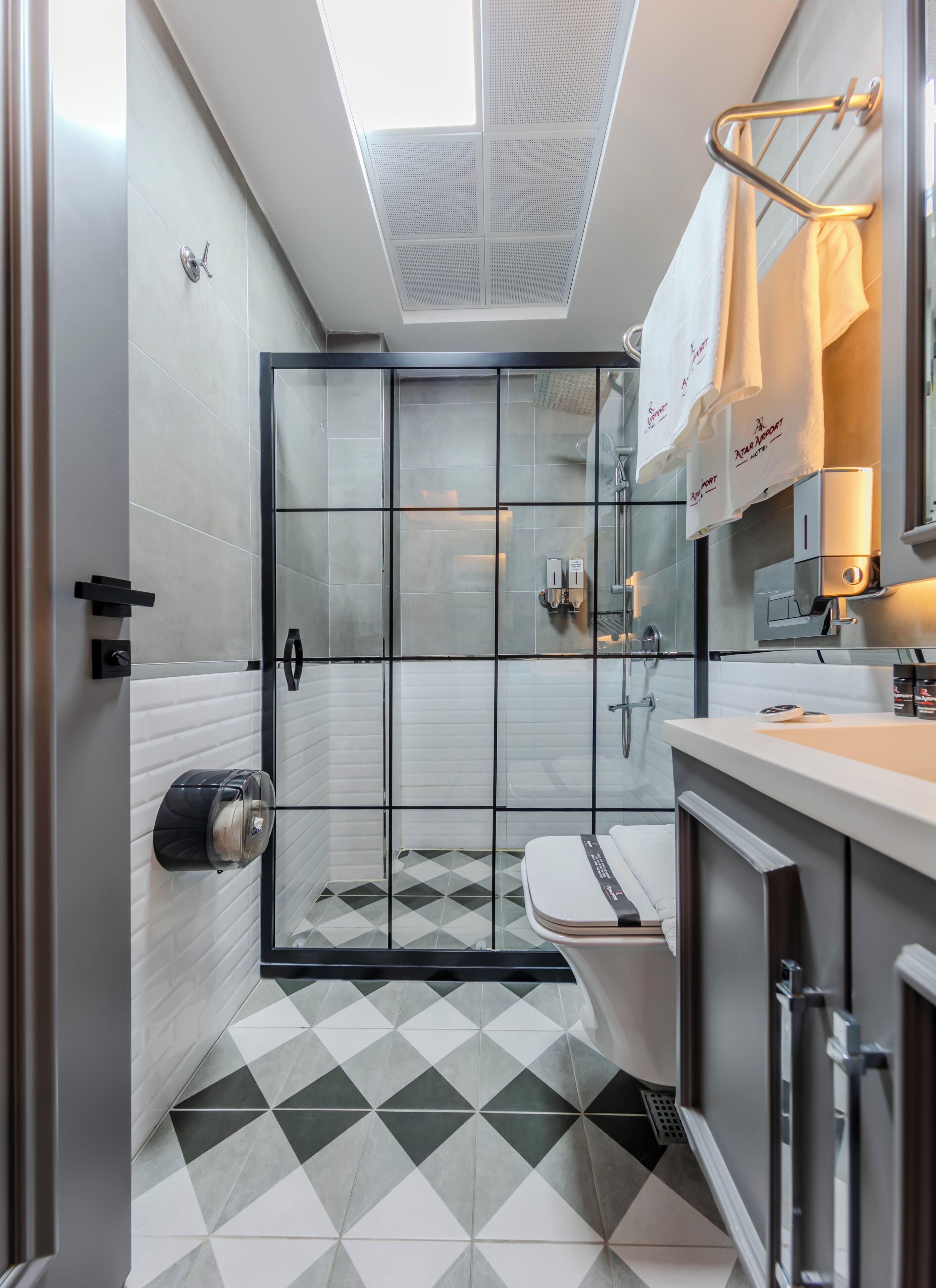 A modern bathroom with a shower, toilet, and sink. It features a geometric patterned floor and a skylight above the shower.