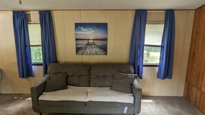 A gray sofa with a beige cushion in a room with wooden walls and blue curtains. - Gateway to keystone lake, Jellystone park , new mannford ramp& the harbor . (Mannford)