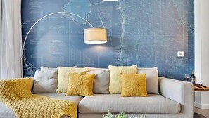 Una sala moderna con un sofá, cojines amarillos, un plaid de punto, una pared con un mapa y una lámpara.