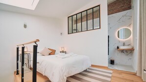 A bedroom with a bed, bedside lamps, a wooden floor, a striped rug, a skylight, a brick wall, a bathroom with a sink and mirror, and a wooden shelf.