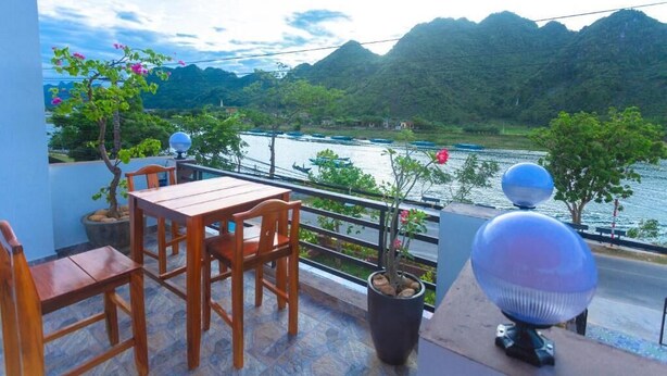 Een balkon met een houten tafel en stoelen, planten in potten, en uitzicht op een rivier en bergen.