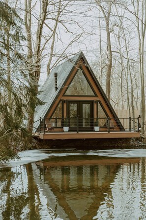 Una cabaña con techo triangular, ventanas grandes y una terraza con vistas a un cuerpo de agua.