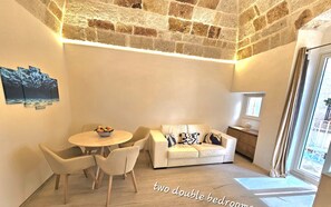 Un appartement comprenant deux chambres avec lits doubles, un plafond en pierre, une table à manger avec des chaises, un canapé avec des coussins et une petite table d’appoint.