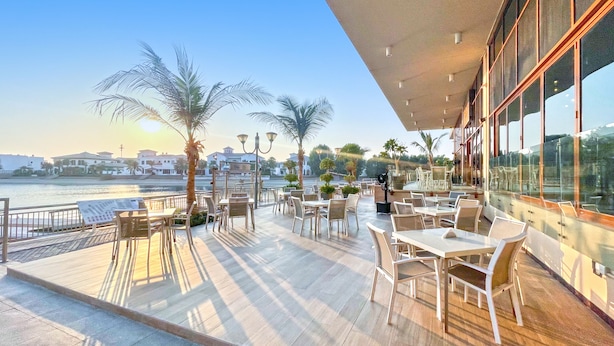 A terrace with tables and chairs overlooking a body of water, palm trees, and a clear sky. - bnbme | Palm Views West | STUDIO (Dubai)
