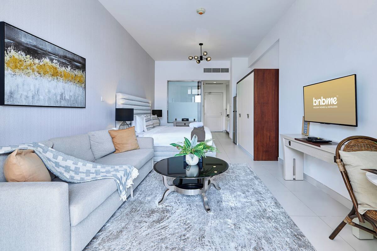 Une chambre d’hôtel moderne avec un canapé gris, une table basse agrémentée d’une plante, un téléviseur fixé au mur et un coin repas avec une chaise en osier.