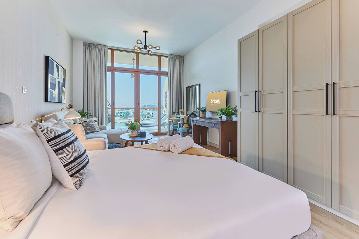 Une chambre d’hôtel avec un grand lit, un bureau, une chaise et une vue sur la ville grâce à de grandes fenêtres.