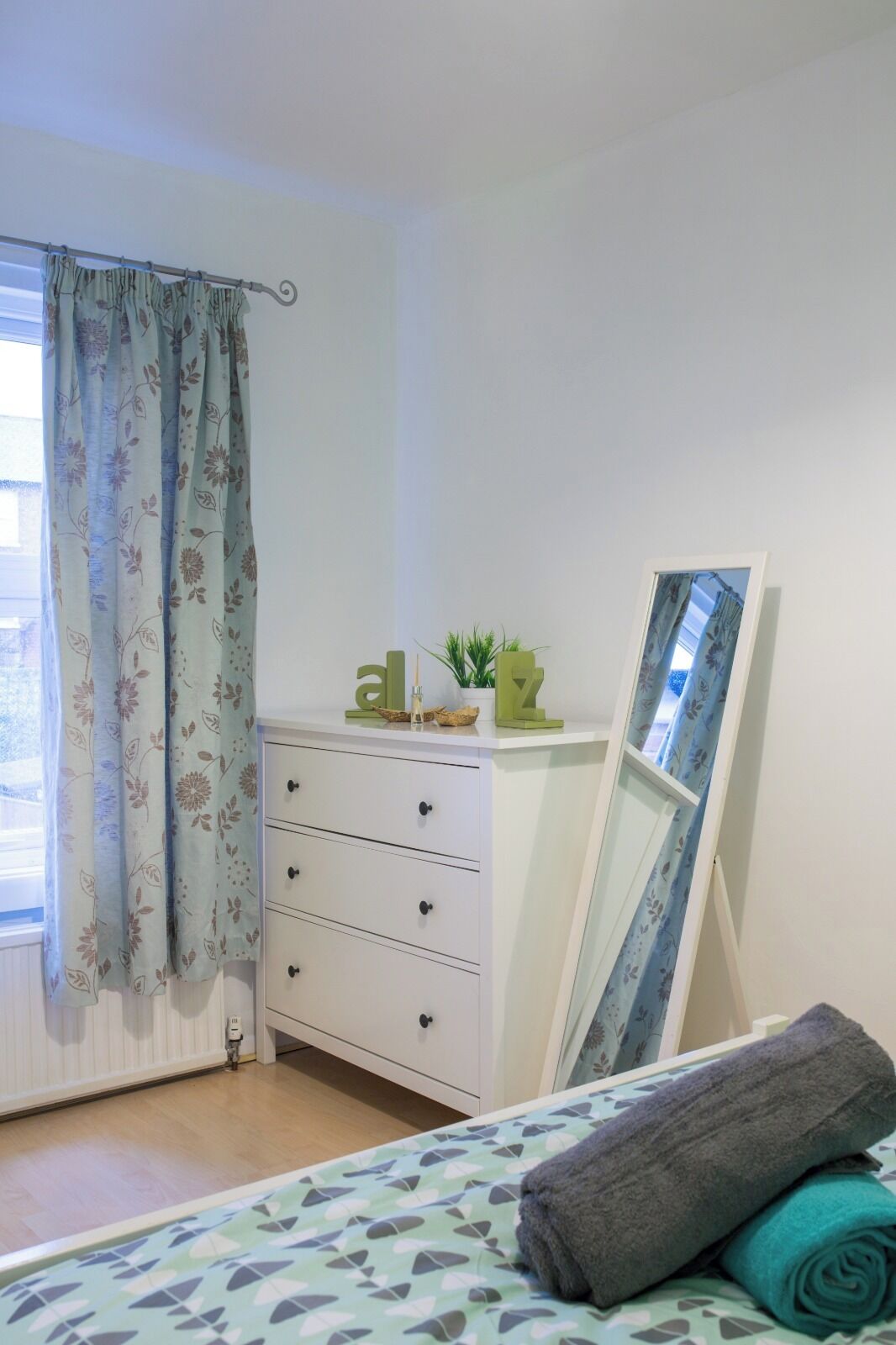 A bedroom with a bed, a dresser, a mirror, and a window with floral curtains.