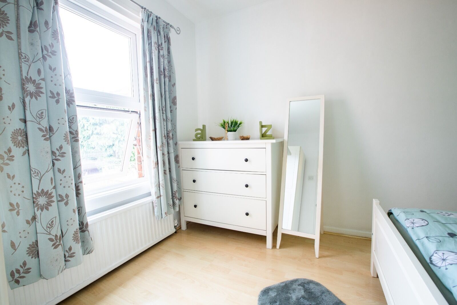 A bedroom with a white dresser, a mirror, a bed with a patterned bedspread, and a window with floral curtains.