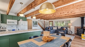 A spacious living area with a wooden ceiling, a kitchen with green cabinets, a dining table with a bowl centerpiece, and a fireplace. - Marina Beach Cabin - Dog-Friendly Home with Deck! by Yosemite Region Resorts (Groveland)