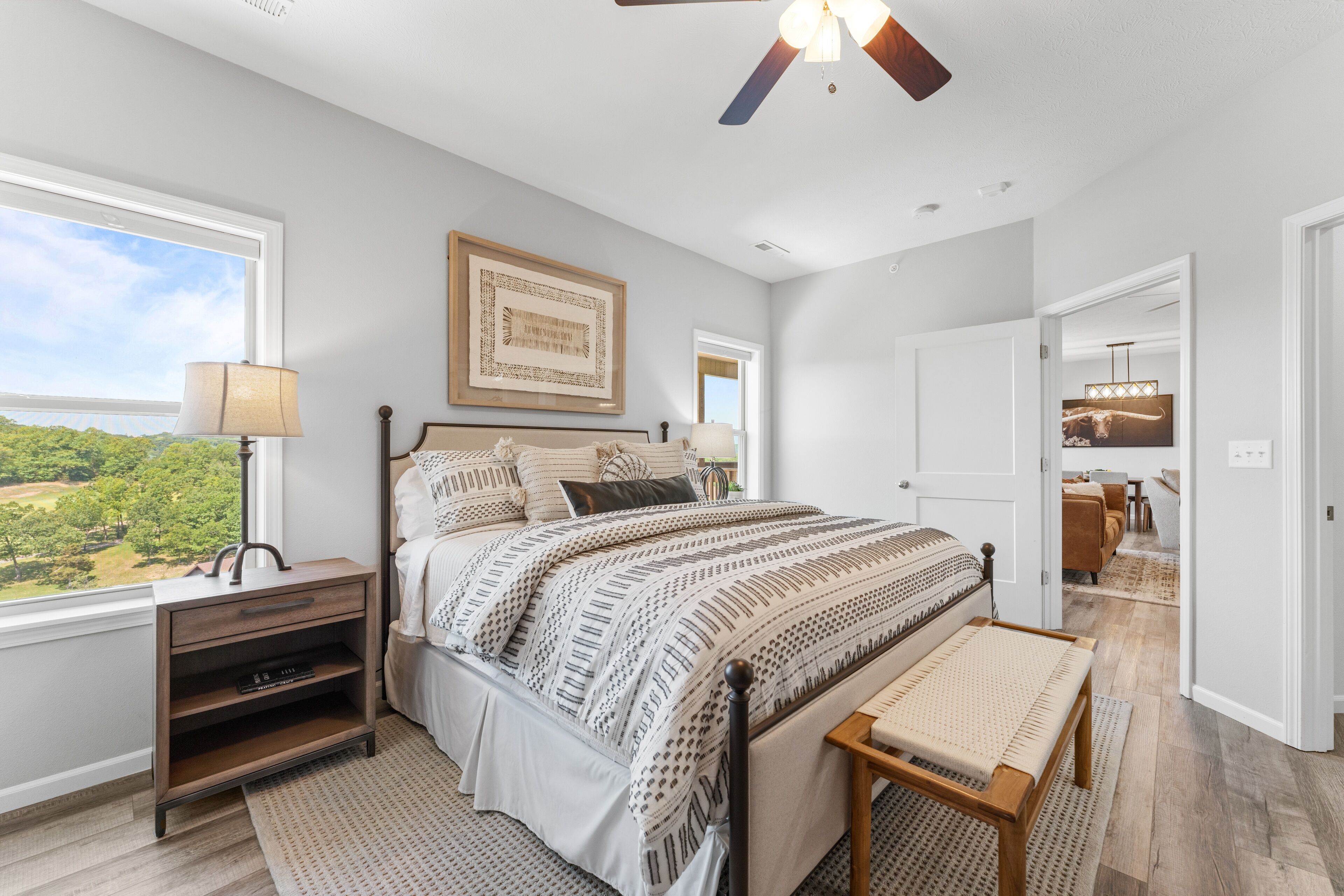 A bedroom with a large bed, bedside tables, a nightstand, a bench, a ceiling fan, and a view of the outdoors.