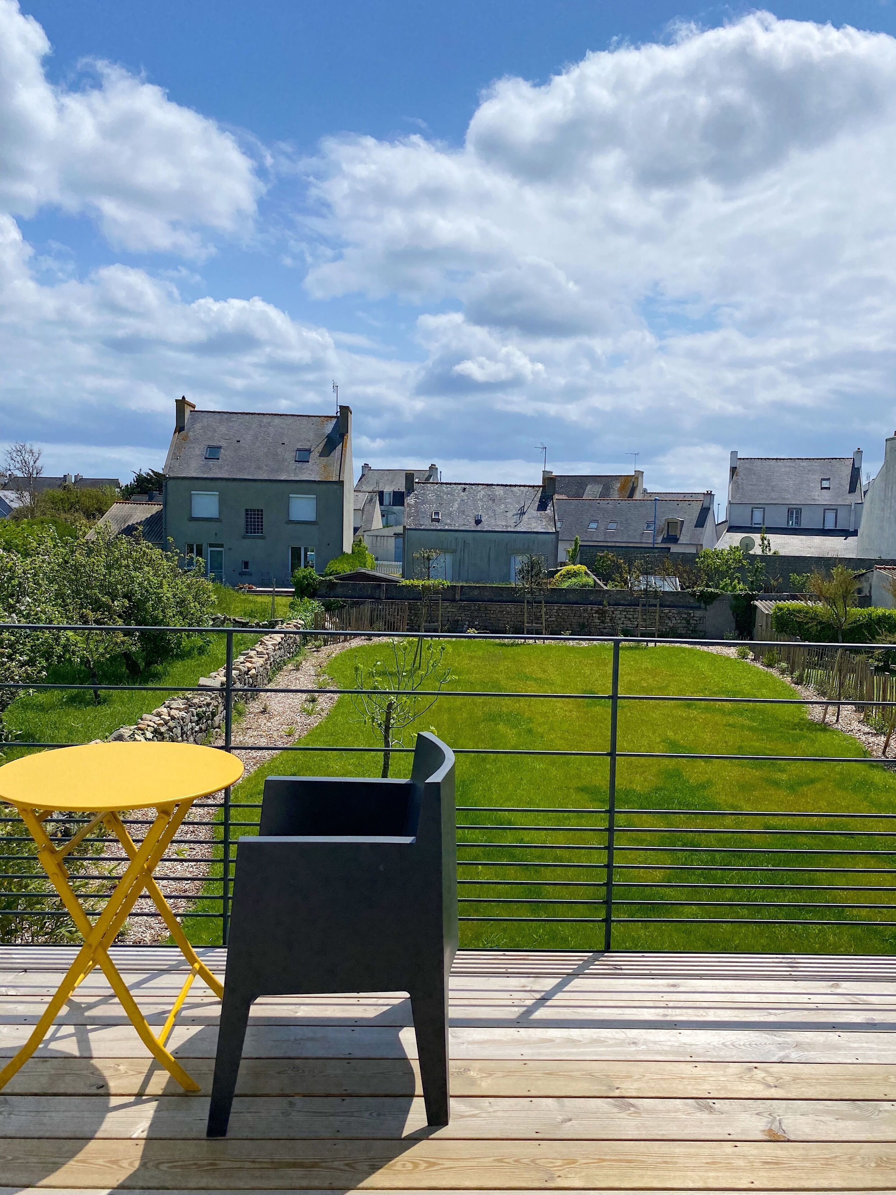 Una terraza con una mesa amarilla y una silla negra, con vistas a un césped cuidado y casas al fondo.