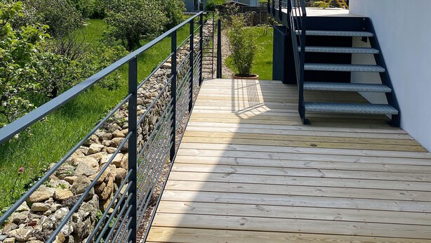 Eine Holzterrasse führt zu einer Treppe mit Metallgeländer, umgeben von Grünpflanzen und einer Steinmauer.