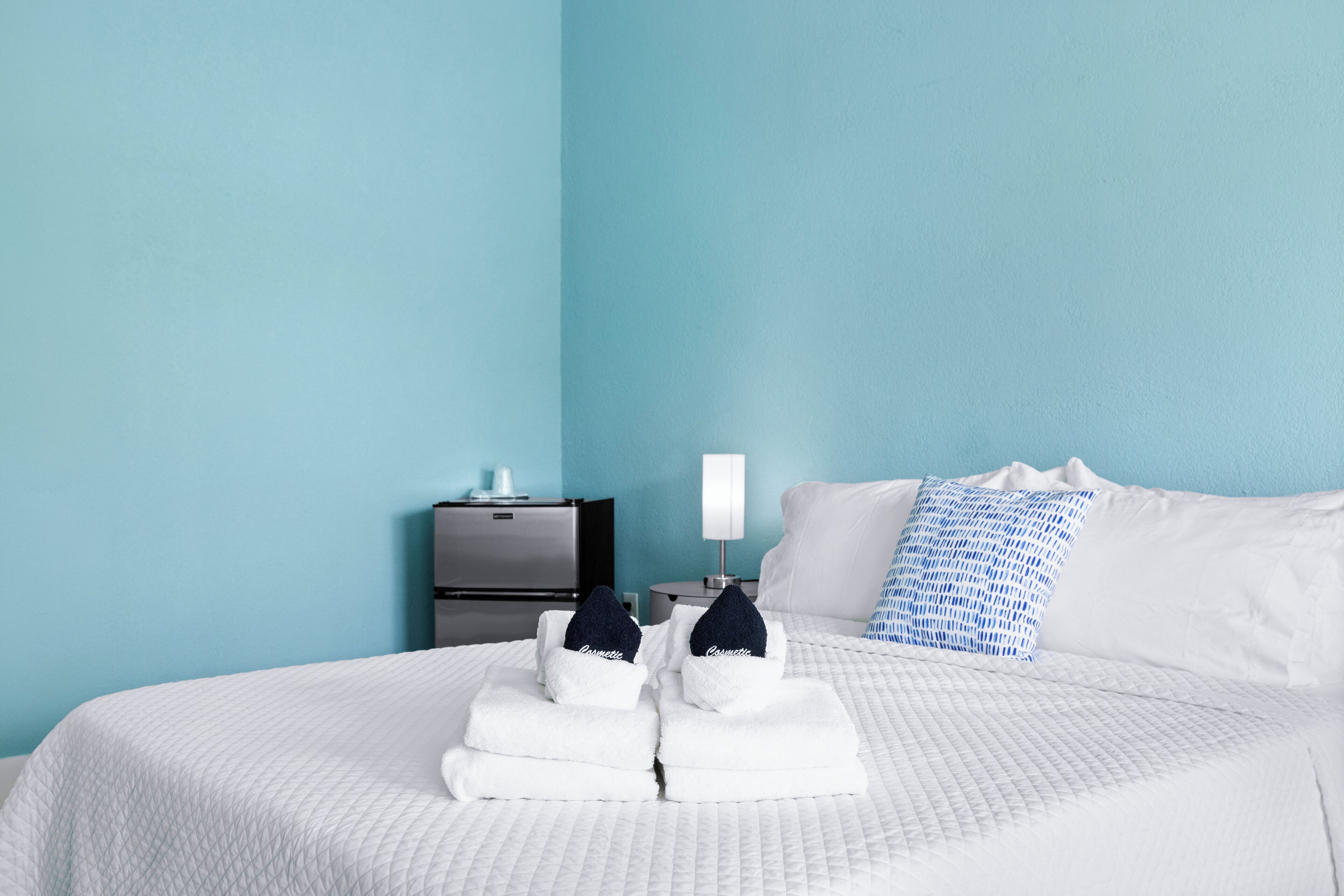 A neatly made bed with folded white towels and a blue pillow, a bedside table with a lamp, and a small cabinet.