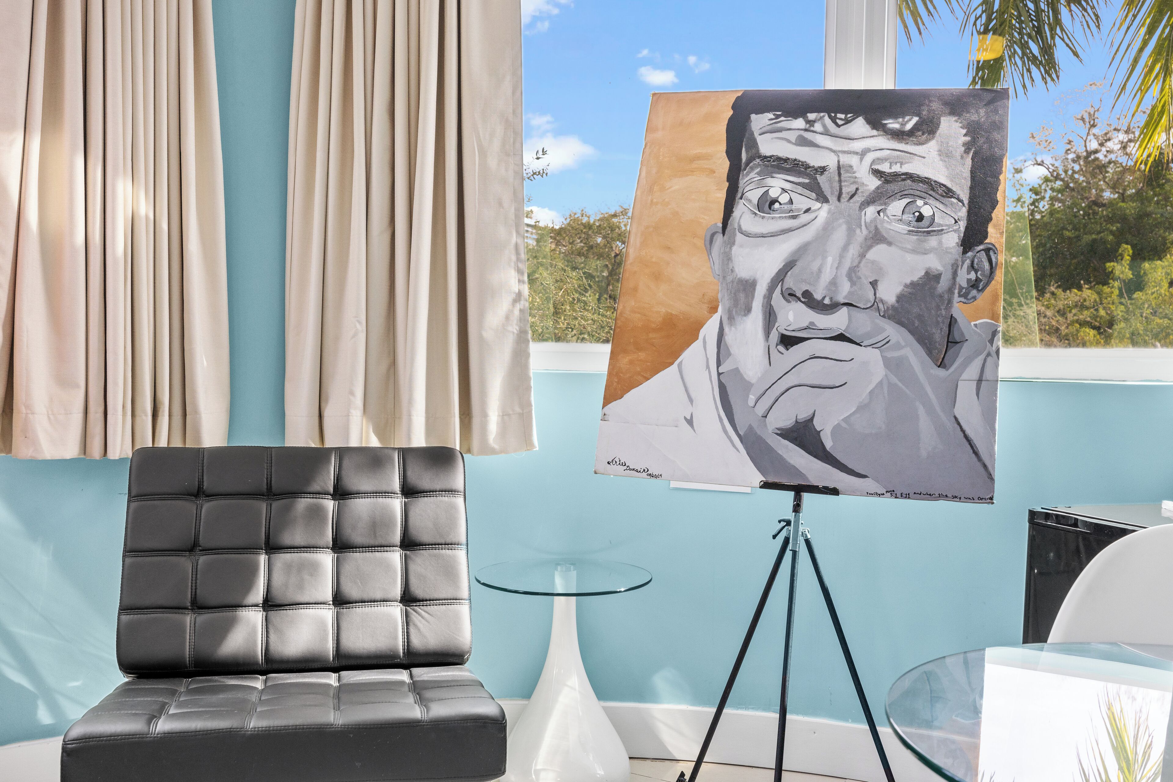A room with a black leather sofa, a glass table, and a painting on a tripod stand.