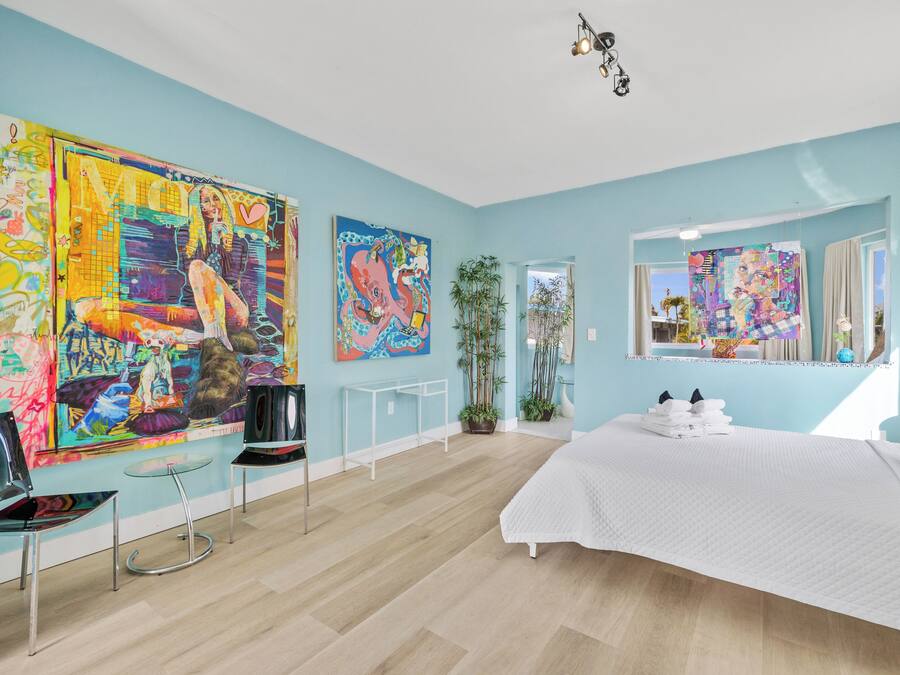 A modern bedroom with a large bed, a seating area with a table, and multiple colorful artworks on the walls.