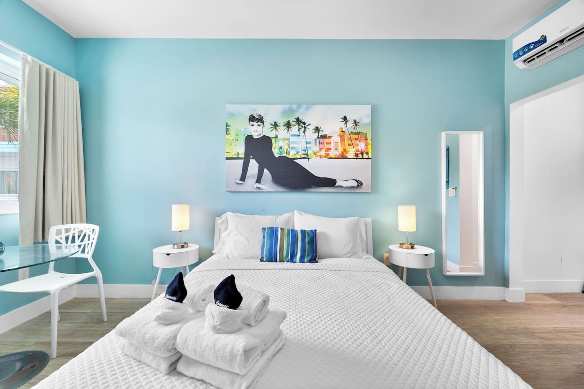 A modern hotel room with a large bed, two bedside tables, a glass table, and a painting of a cityscape.