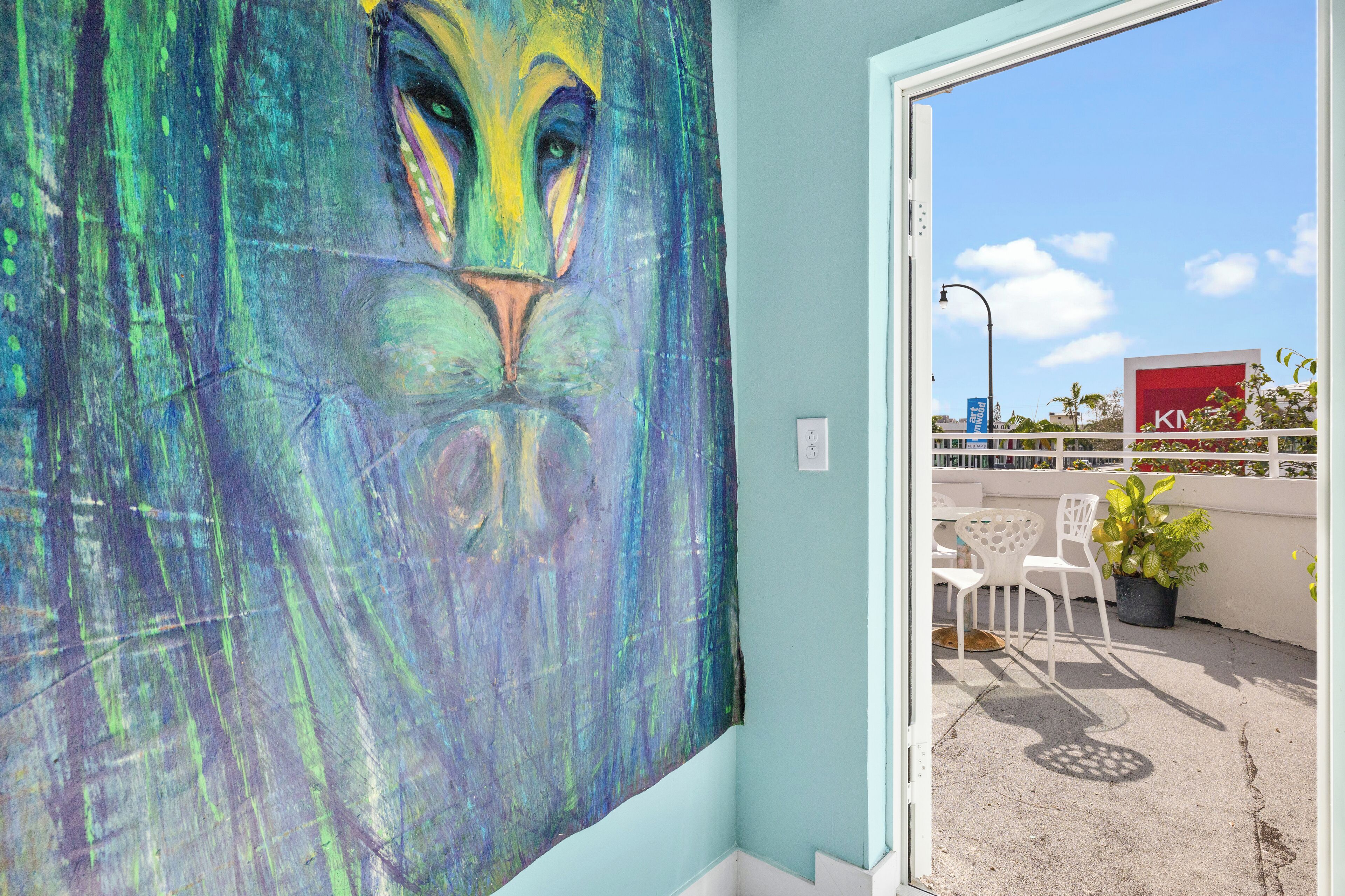 A room with a large abstract painting on the wall, a window with a view of a building, and a balcony with chairs and a potted plant.