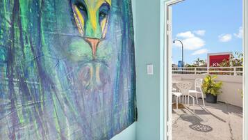 A room with a large abstract painting on the wall, a window with a view of a building, and a balcony with chairs and a potted plant.