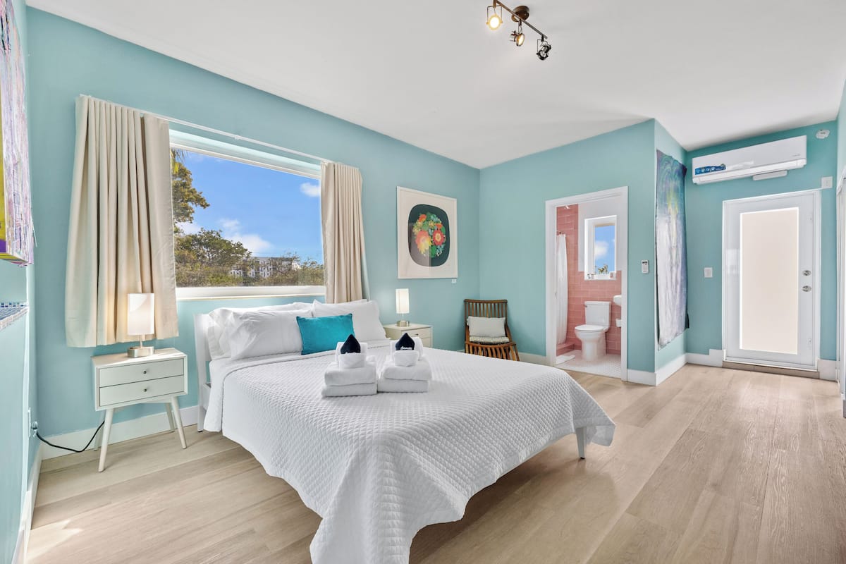 A bedroom with a large bed, a window, a nightstand, a bedside table, a painting, a bathroom with a toilet, and an air conditioning unit.