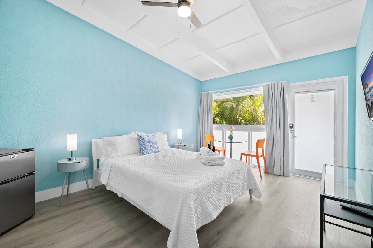 A modern bedroom with a bed, a small table, a chair, a ceiling fan, and a door leading to a balcony.