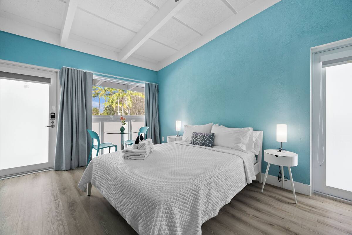 A bedroom with a bed, a nightstand, a window with curtains, a chair, and a view of palm trees.