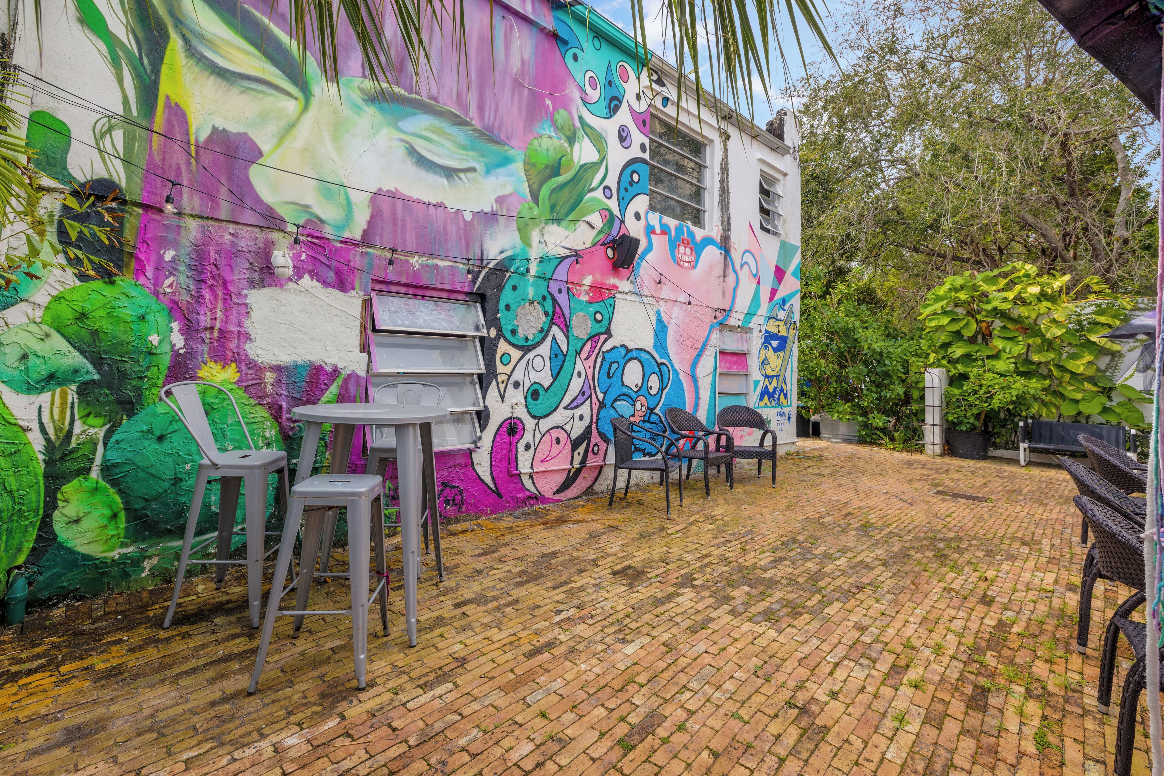An outdoor patio area with a colorful mural on the wall, a brick floor, and several chairs and tables.