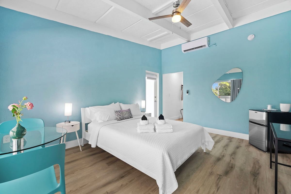 A hotel room with a bed, a desk, a chair, a small table, a mirror, and a vase with flowers.