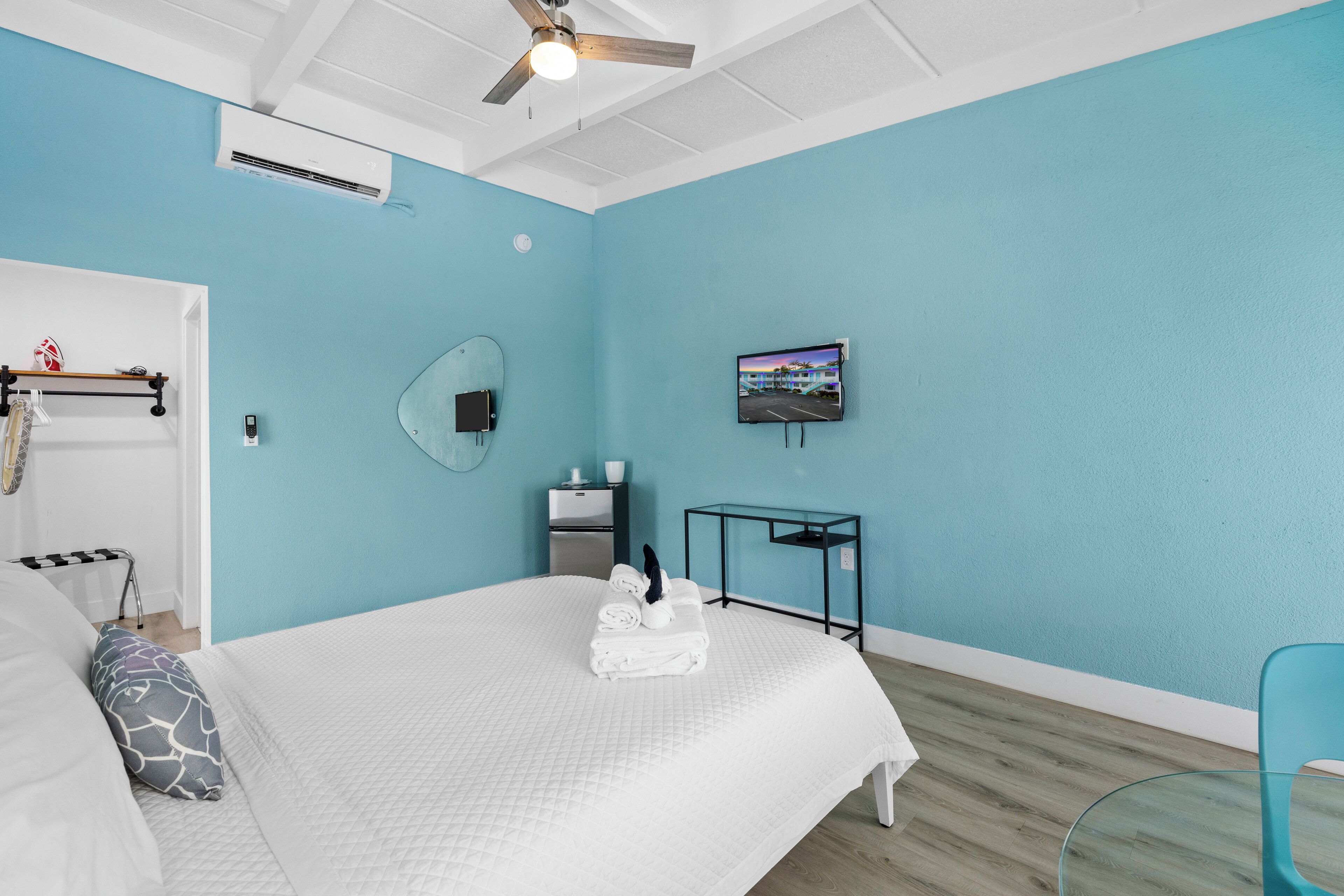 A modern hotel room with a large bed, a ceiling fan, an air conditioning unit, a flat-screen TV, a small table, and a glass chair.