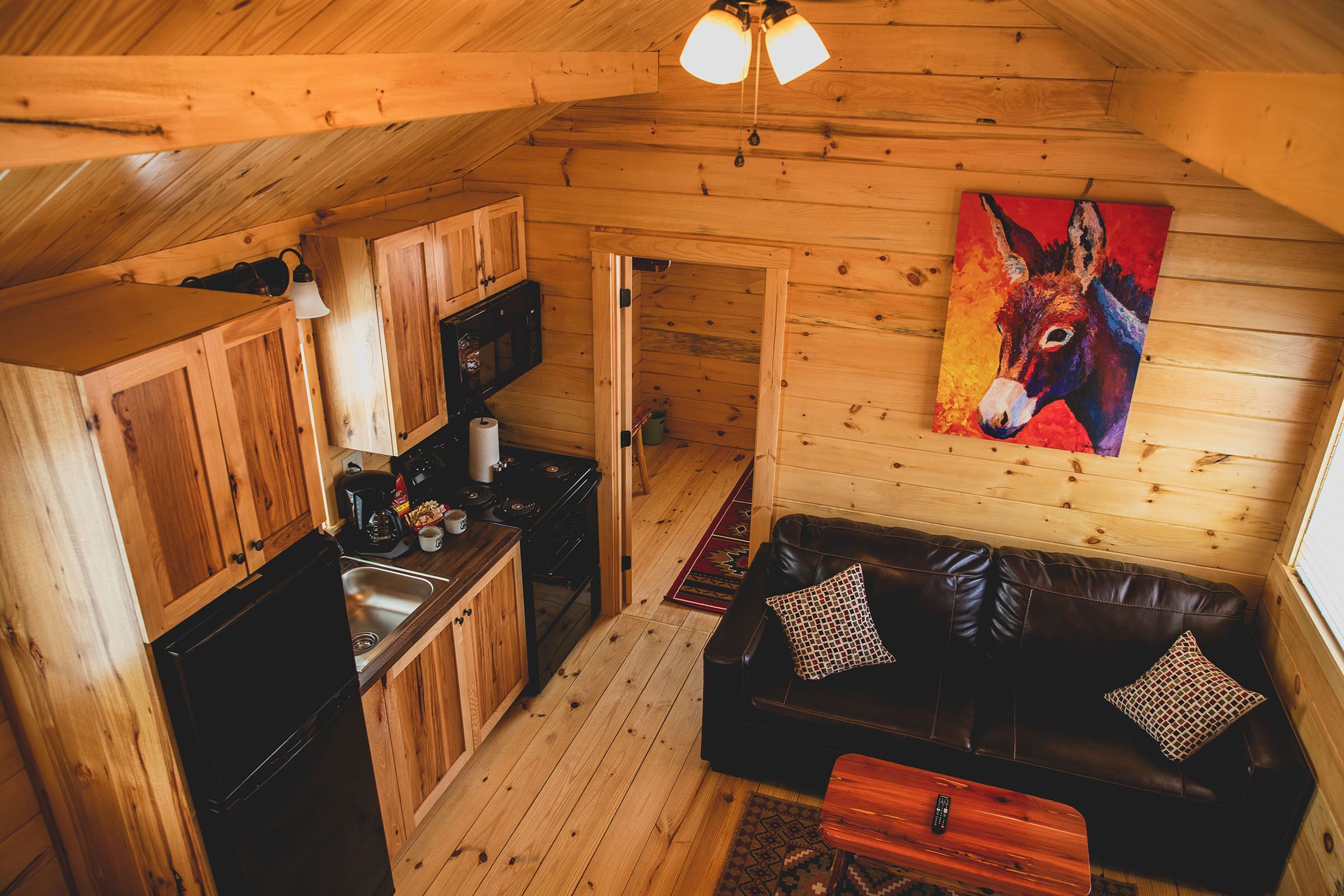 A cozy cabin interior with wooden walls, a black refrigerator, a wooden dining table, a black leather couch, and a colorful donkey painting on the wall.