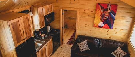 A cozy cabin interior with wooden walls, a black refrigerator, a wooden dining table, a black leather couch, and a colorful donkey painting on the wall.