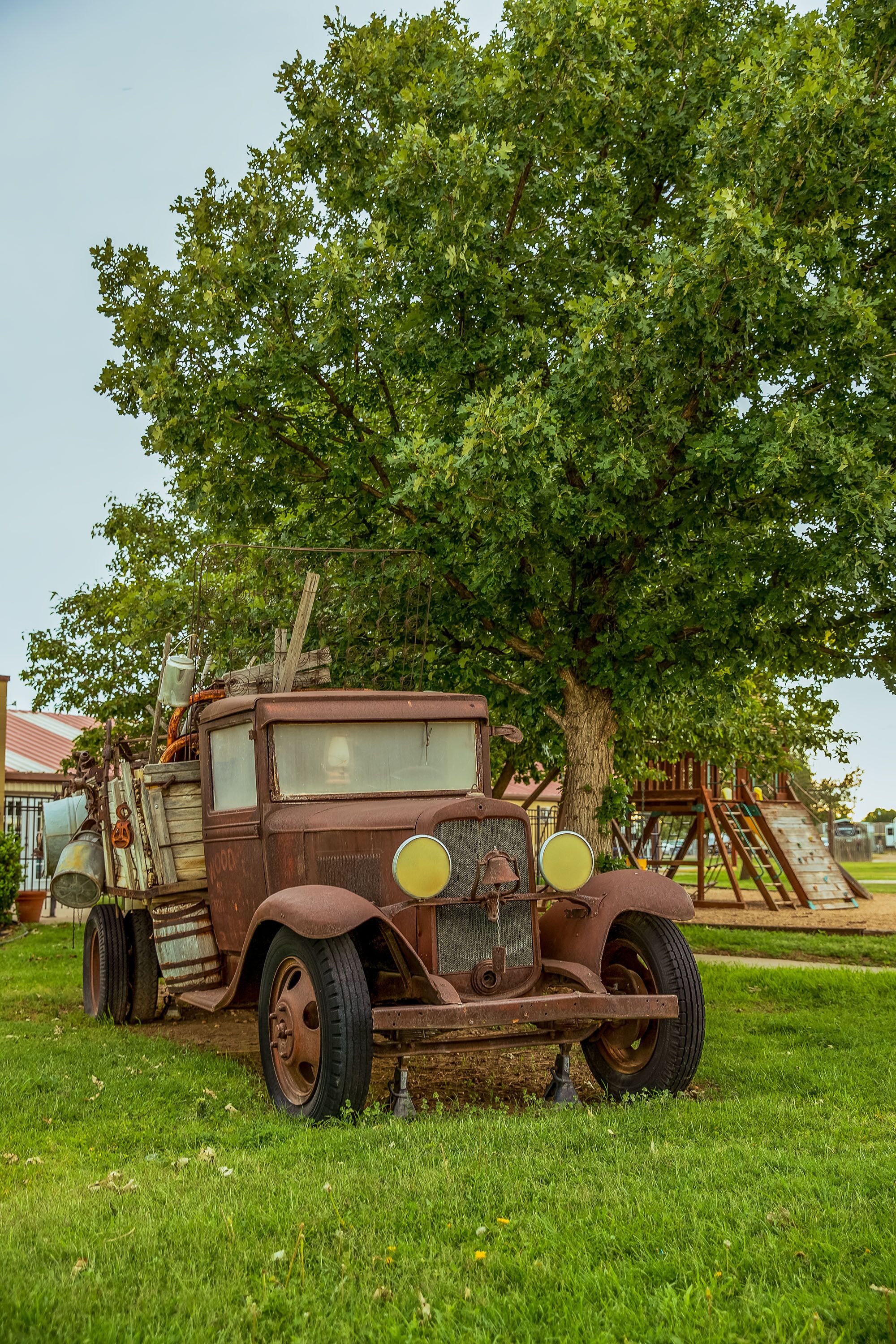 An old, rusted truck parked in a grassy area with a large tree in the background.