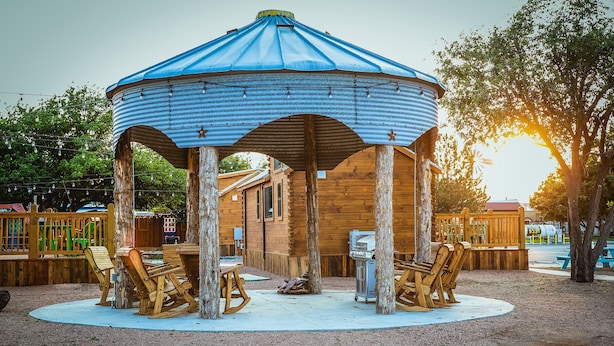 A wooden gazebo with a blue metal roof, surrounded by wooden benches and a grill. - The Big Texan - Cabins & Wagons  (Amarillo)