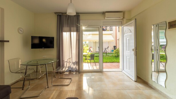 A modern living room with a glass table, chairs, a flat-screen TV, and a sliding glass door leading to an outdoor patio. - Nano's Vacation In Nea Skioni  (Kassandra)
