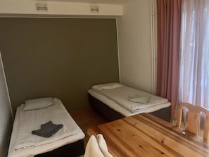 Ein kleines Zimmer mit zwei Einzelbetten, einem Holztisch und einem Fenster mit Vorhängen.