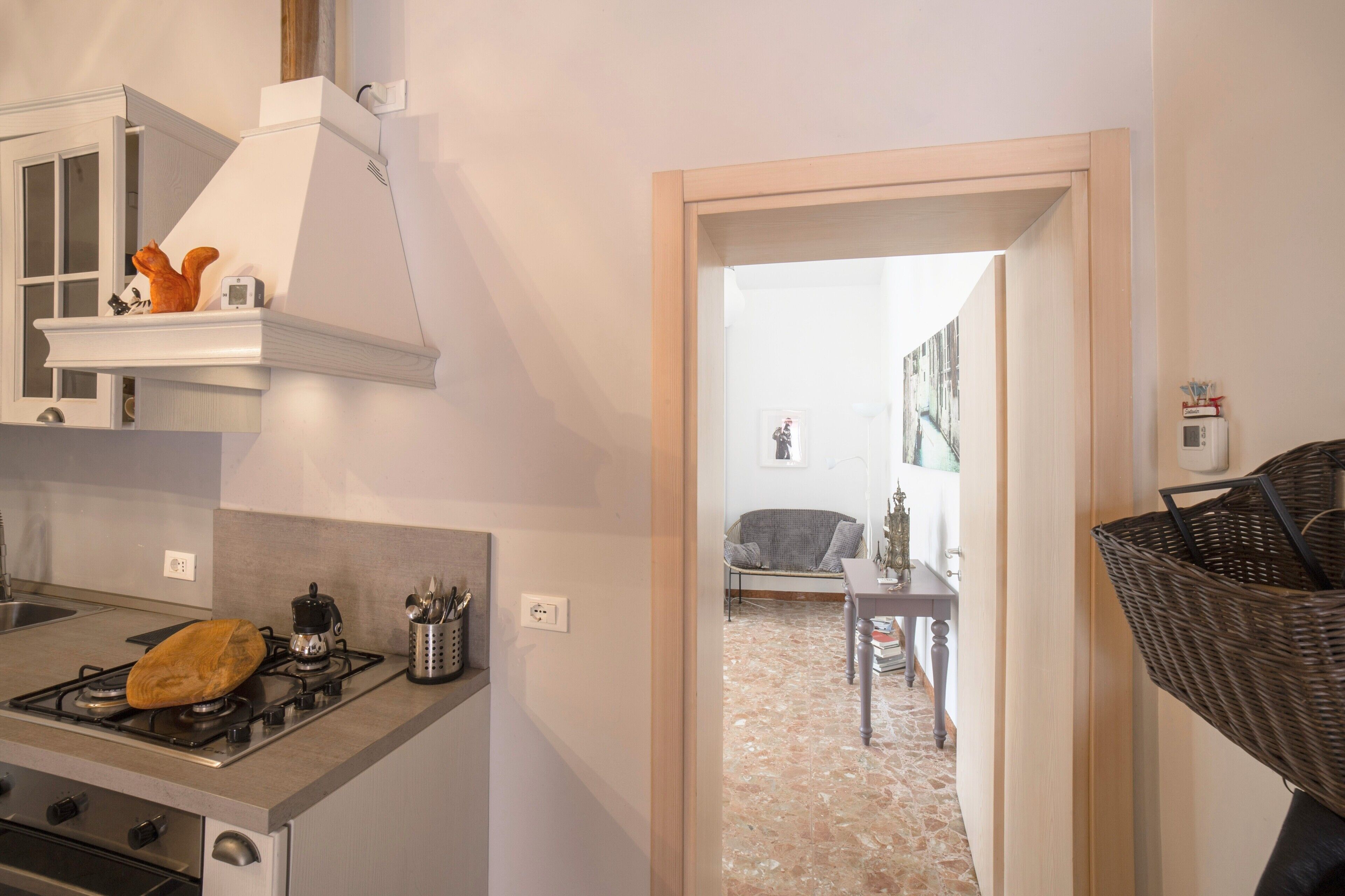 A modern kitchen with a gas stove, oven, and a white range hood. There is a dining area visible through an open doorway with a sofa and a table.