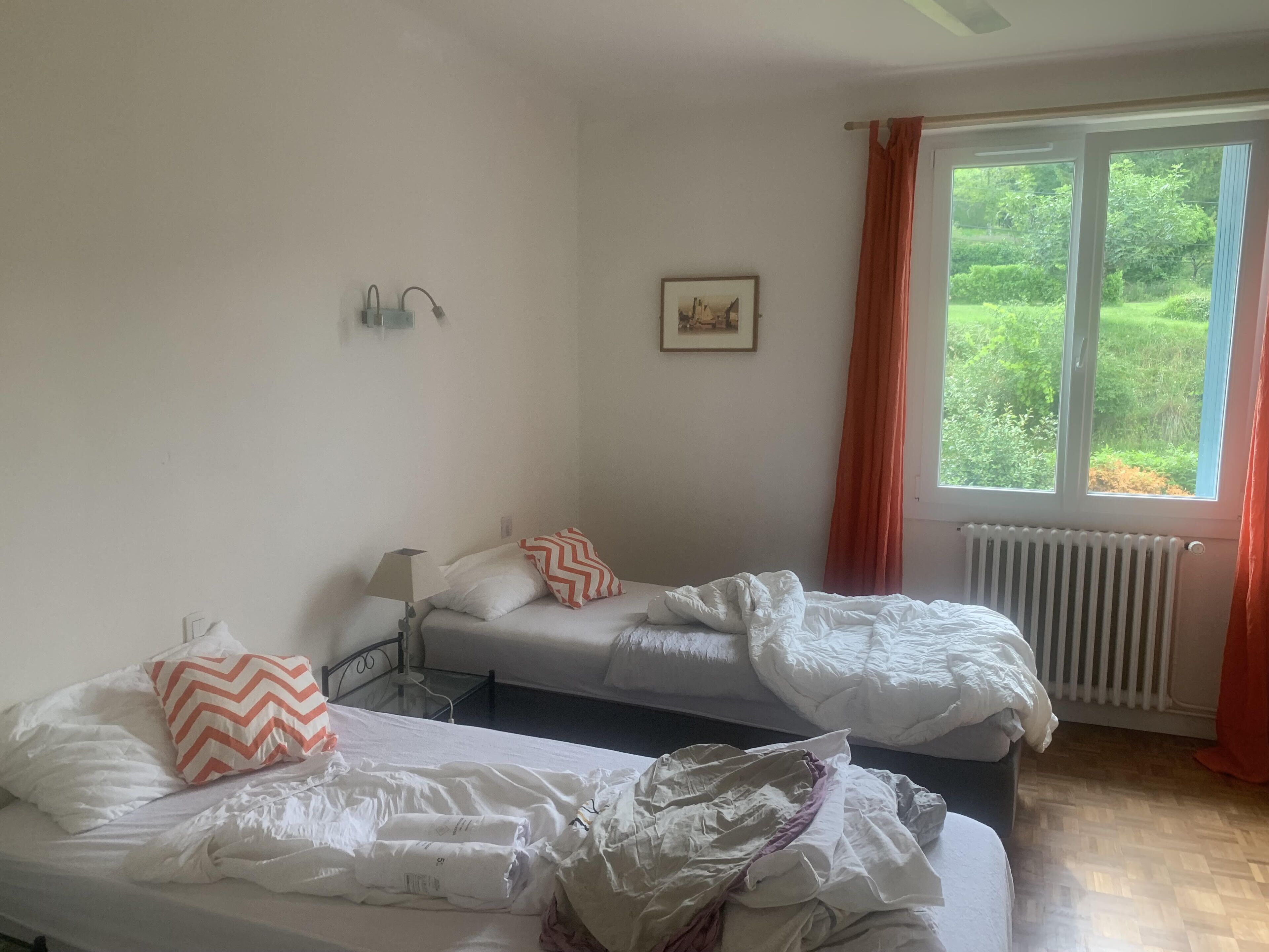 Una camera d'albergo con due letti singoli, un tavolo, una lampada, un quadro appeso al muro e una finestra con tende.