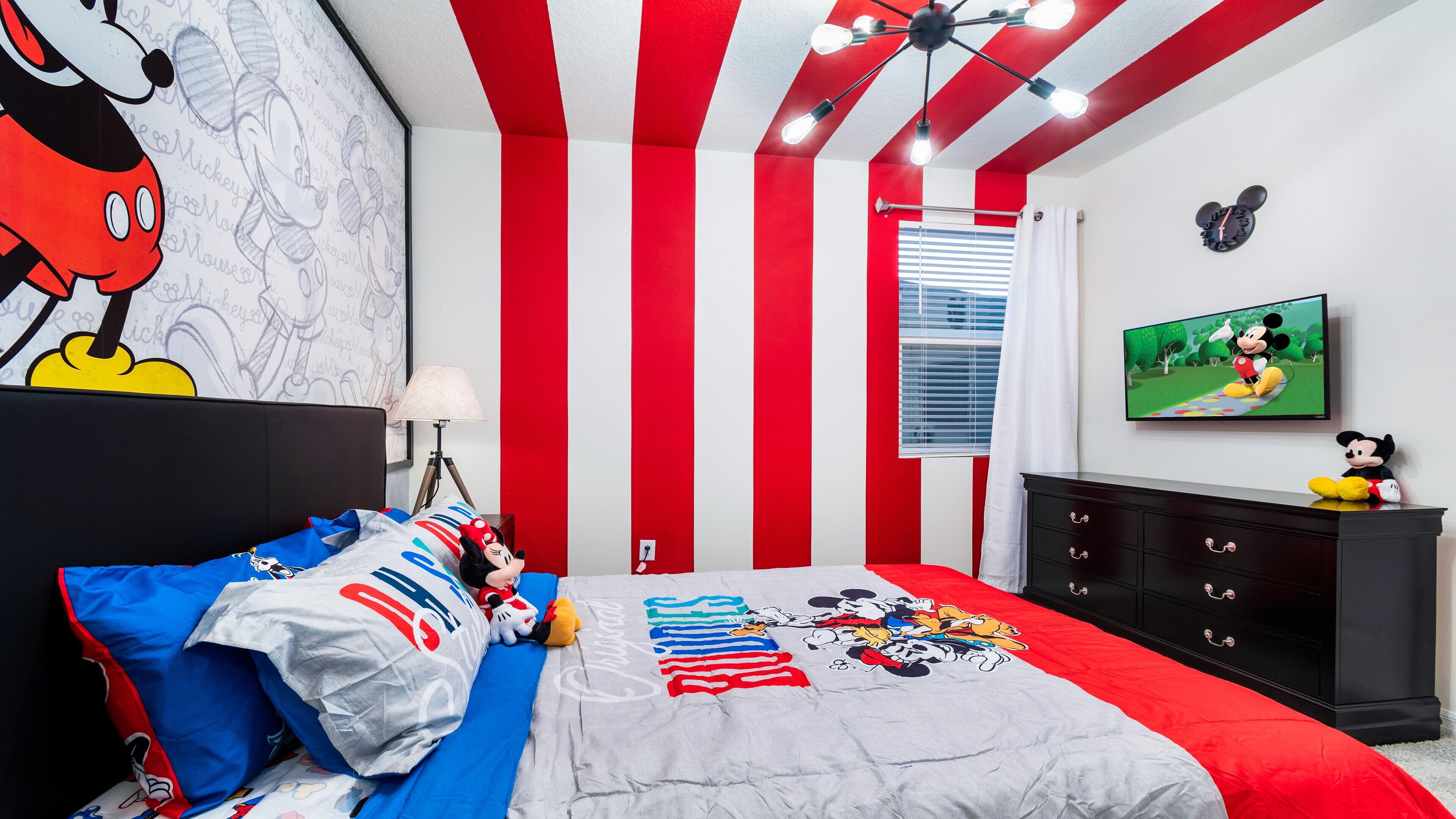 A bedroom with a Mickey Mouse mural, striped red and white walls, a bed with Mickey-themed bedding, a TV, and a dresser.