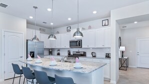 A modern kitchen with white cabinets, a granite island with bar stools, stainless steel appliances, pendant lighting, and a small side table with a lamp. - Last Minute Disney Getaway! Private Pool & Themed Bedrooms. Book Now! #59697 (Orlando)
