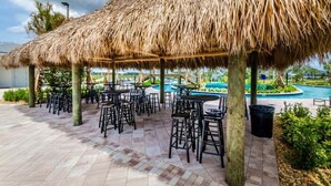 A covered outdoor seating area with tables and chairs, a thatched roof, and a pool in the background. - Last Minute Disney! Private Pool, Hot Tub, Game Room & Themed Bedrooms. Book Now! #57281 (Orlando)