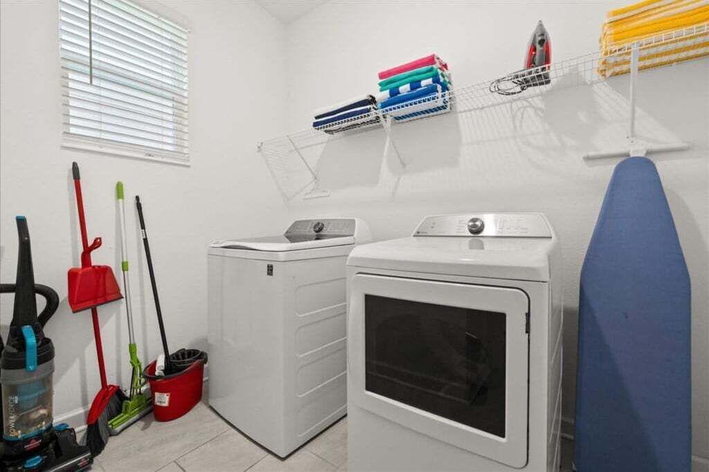 A laundry room with a washing machine, dryer, ironing board, and various cleaning tools.