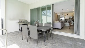 Un patio exterior con una mesa y sillas de comedor, una parrilla y una puerta corrediza de vidrio que da a un espacio interior con sofá y electrodomésticos de cocina.